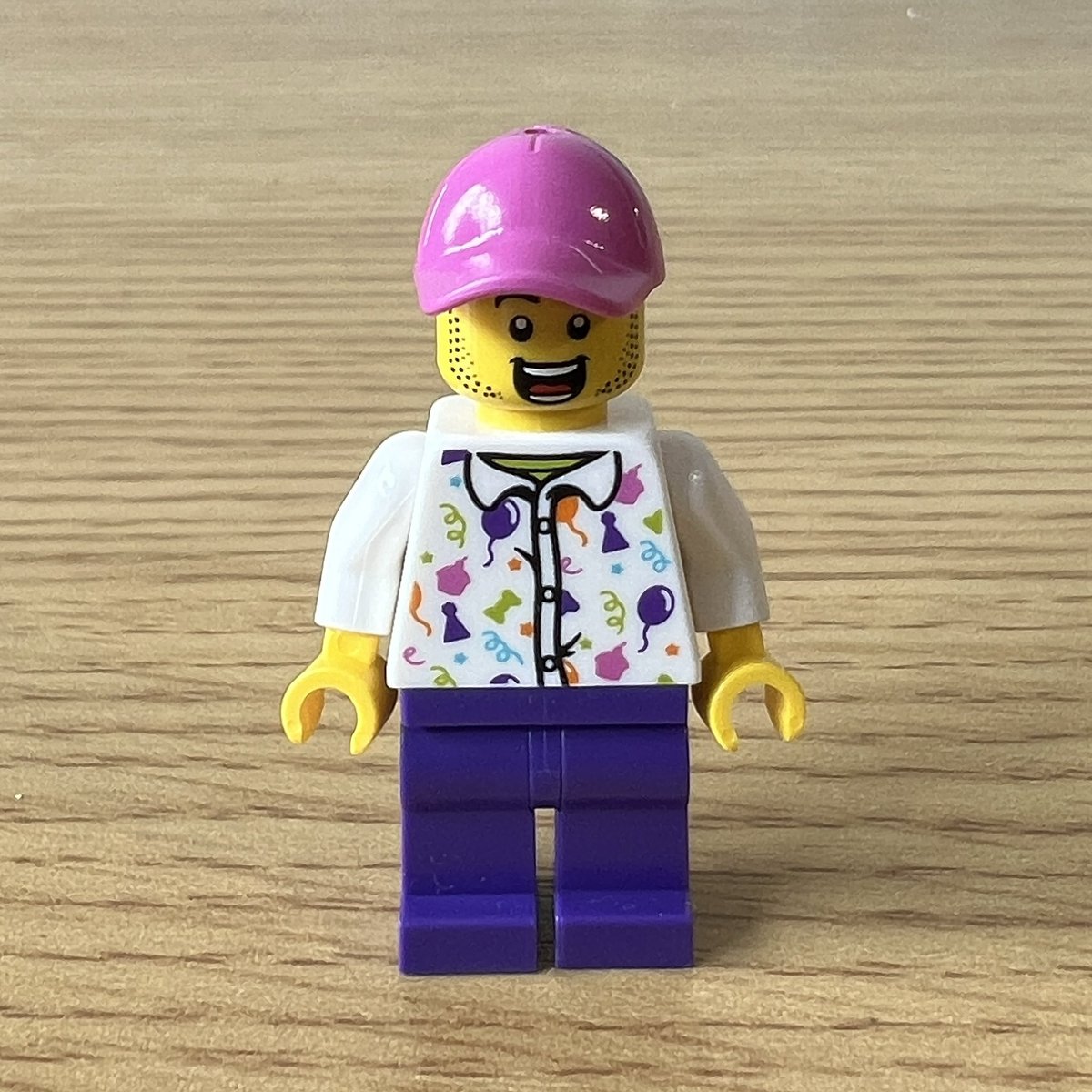 A closeup of a lego minifigure wearing a white shirt with pictures of balloons and candy floss. They’re also wearing purple trousers and a pink baseball cap.