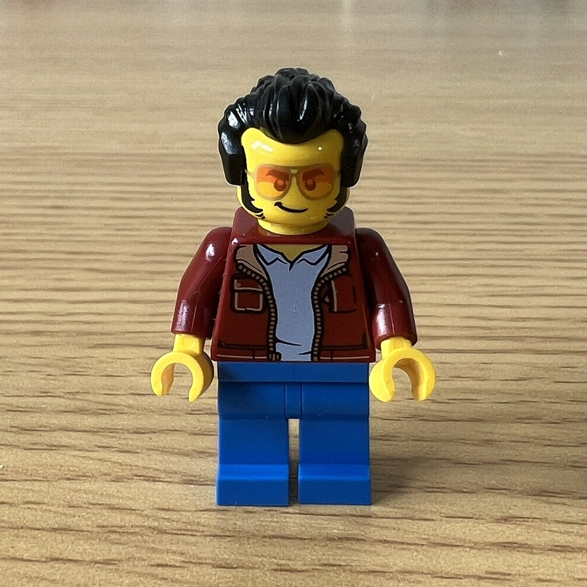 A closeup of a lego minifigure with slicked back hair and sideburns. He has orange tinted glasses, a red leather jacket, and jeans.