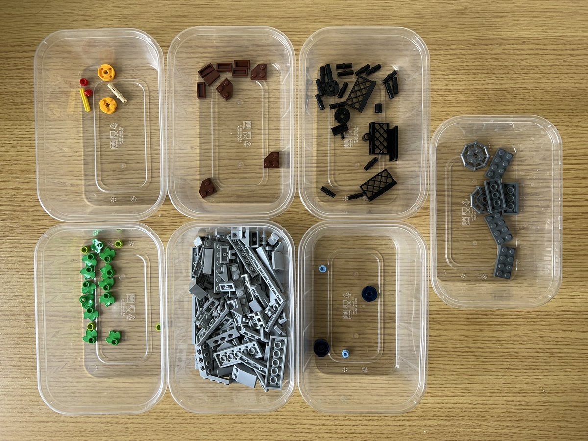 Seven tubs filled with lego sorted by colour. Most of the pieces are light grey, but there’s a tub with a handful of green leaves. The tub of blue pieces only has four, one of which is a cylinder used as a support for a roller coaster.