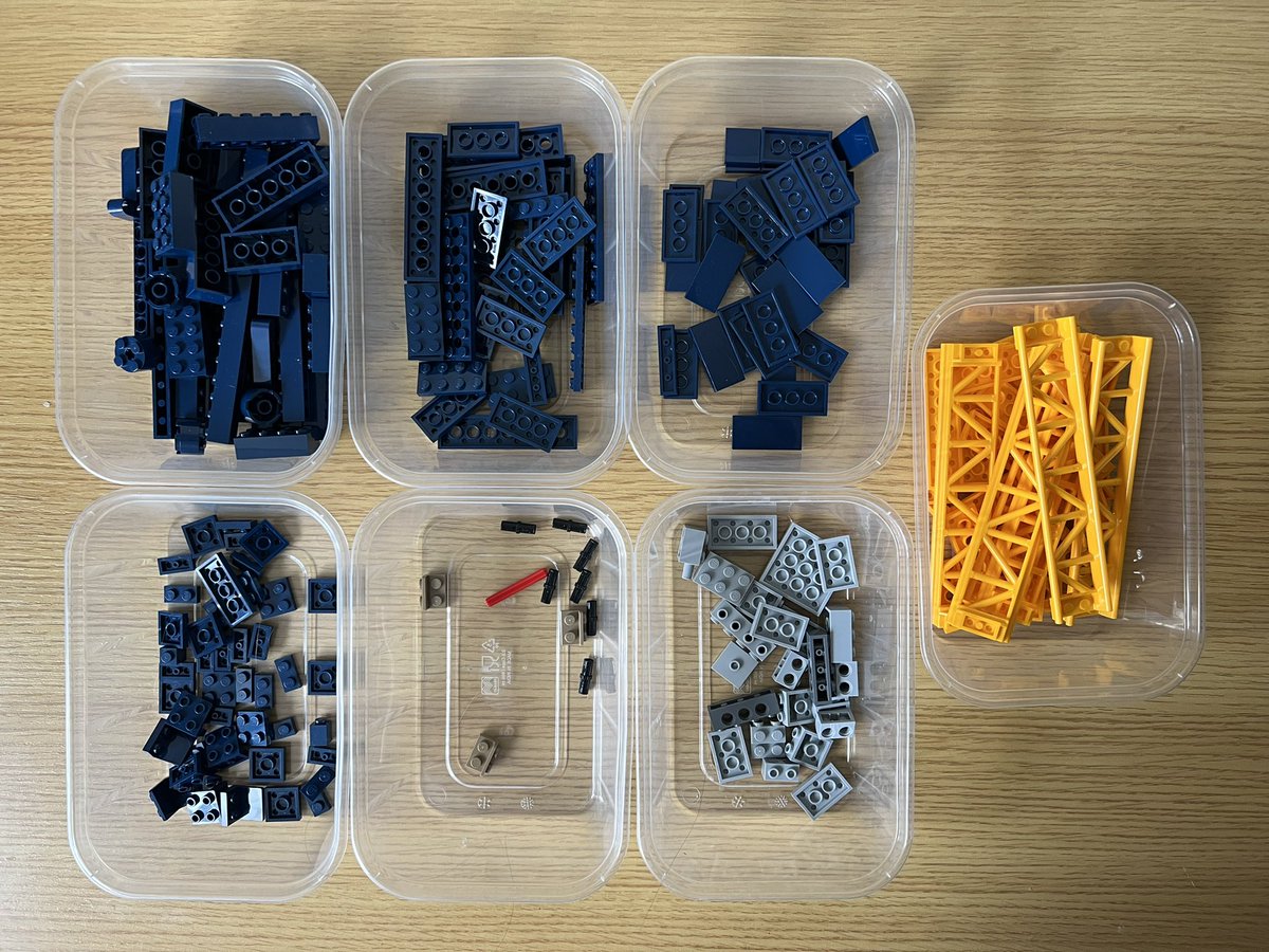 Seven tubs of lego. Four tubs contain dark blue pieces, one has grey pieces, one has yellow pieces including roller coaster track, and the last one has black and brown pieces.