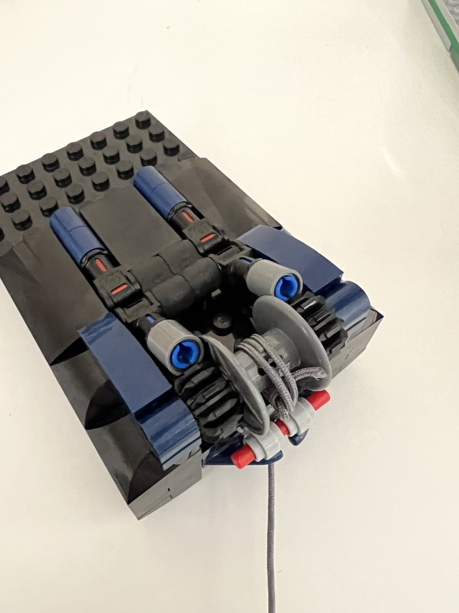 A lego structure with string wrapped around a spool attached to two gears. There are grey pegs resting on the gears so they can only turn in one direction.