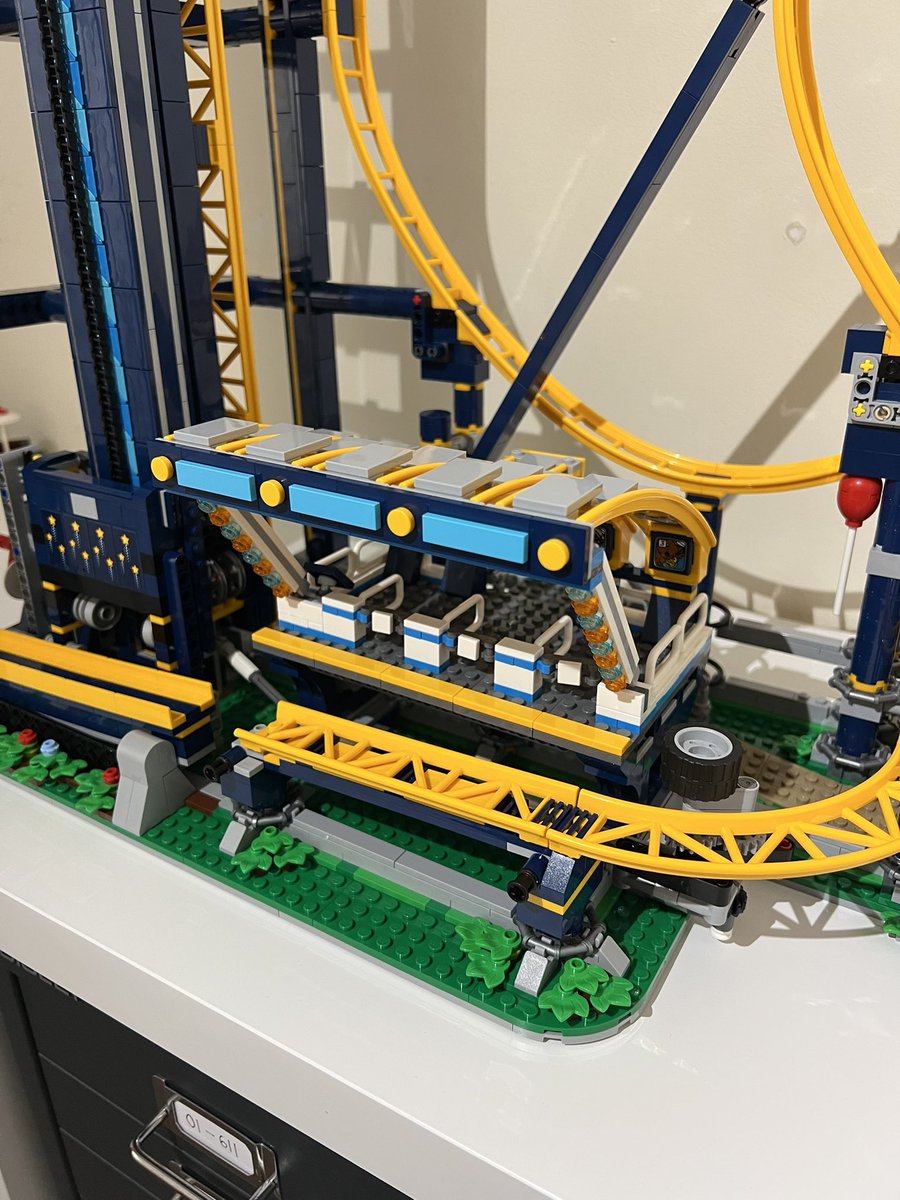 A lego roller coaster station. It has a curved yellow roof with grey tiles, a blue striped design on the front, and blue and orange lights down the supports.